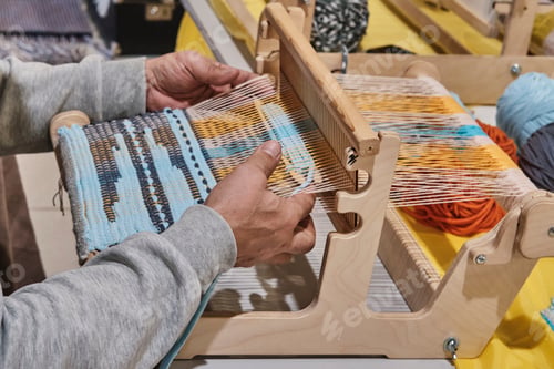 Preview: Hands of senior man weaving small rug with pattern on manual table loom.