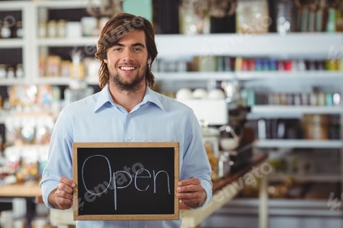 Preview: Adult man standing inside cafe holding chalkboard sign while showcasing espresso machine and jars
