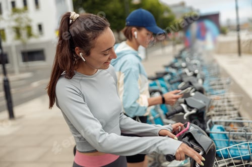 Preview: Two young female friends renting bicycle in bike sharing city service for walking
