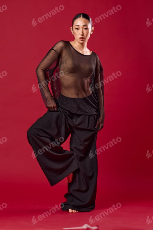 Preview: Fashion asian woman in black pants and sheer top posing confidently on red background