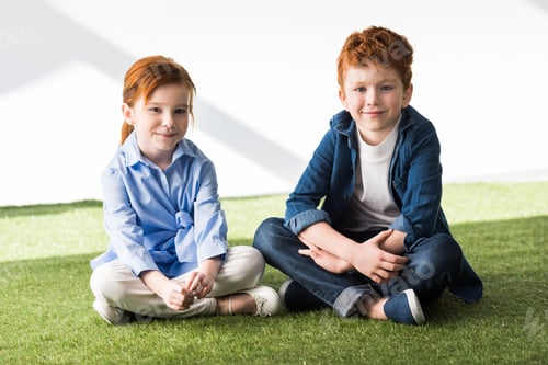 Preview: cute happy redhead children sitting together on grass and smiling at camera on grey