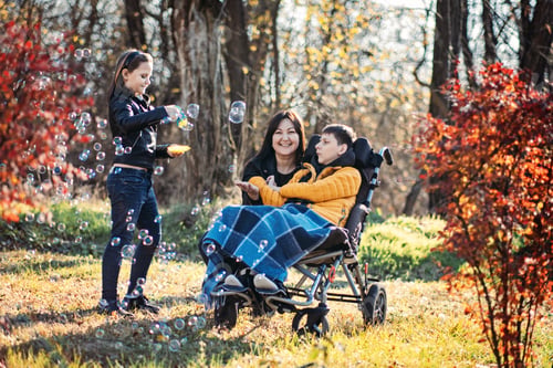 Preview: Mother and sibling playing with bubbles with child in wheelchair, inclusive outdoor play