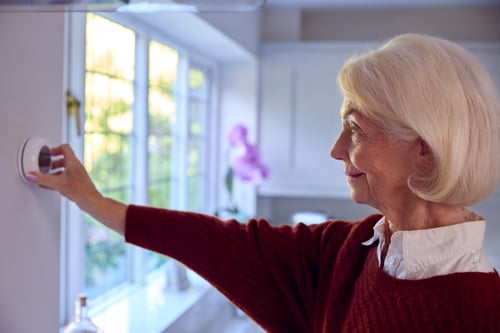 Senior Woman At Home Turning Down Central Heating Thermostat During Cost Of Living Energy Crisis