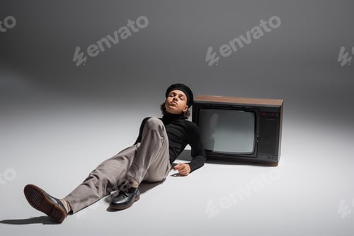 Preview: full length of young african american man with closed eyes lying in sweater and pants near retro tv