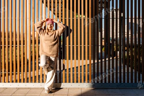 Preview: Fashionable woman posing against a modern wooden fence in a city environment, enjoying the sunlight