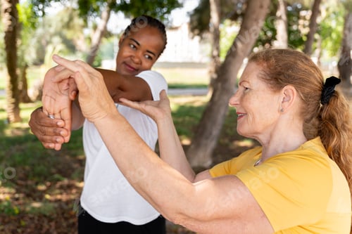 Preview: Diverse senior women exercising in park, one helping another stretch during workout