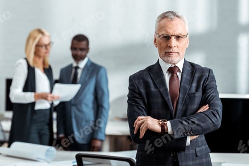 Preview: selective focus of confident businessman in glasses and looking at camera while standing with