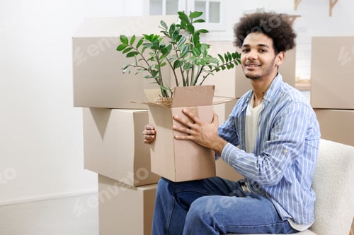 Preview: happy young African American man moves into new home. person is sitting among cardboard boxes