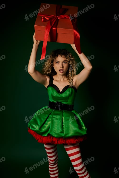 Preview: amazed new year elf in green dress posing with huge present above head and looking at camera