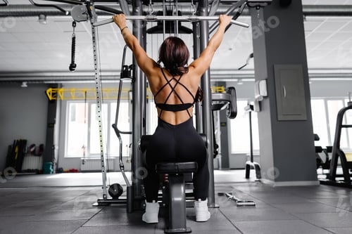 Preview: Female bodybuilder in black suit pulled on sports simulator in gym. Back muscles are very tense.