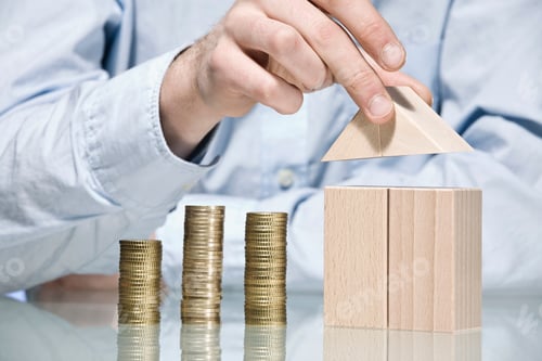 Preview: Person building house with building bricks beside stack of coins, close up
