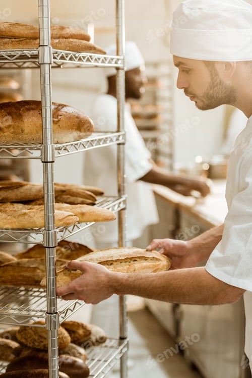 Preview: handsome baker putting fresh bread loaf on shelf at baking manufacture
