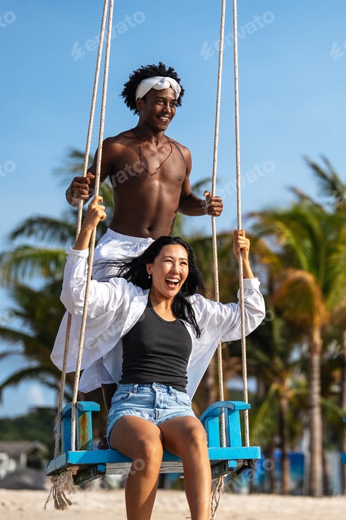 Preview: Happy interracial couple enjoying a fun beach day, swinging under palm trees on a tropical vacation