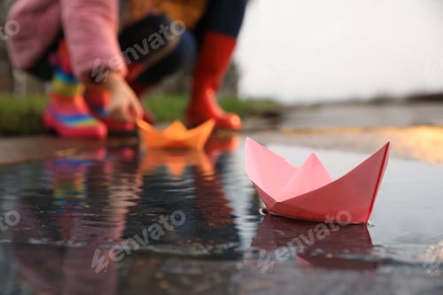 Preview: Little girl and her mother playing near puddle outdoors, focus on paper boat