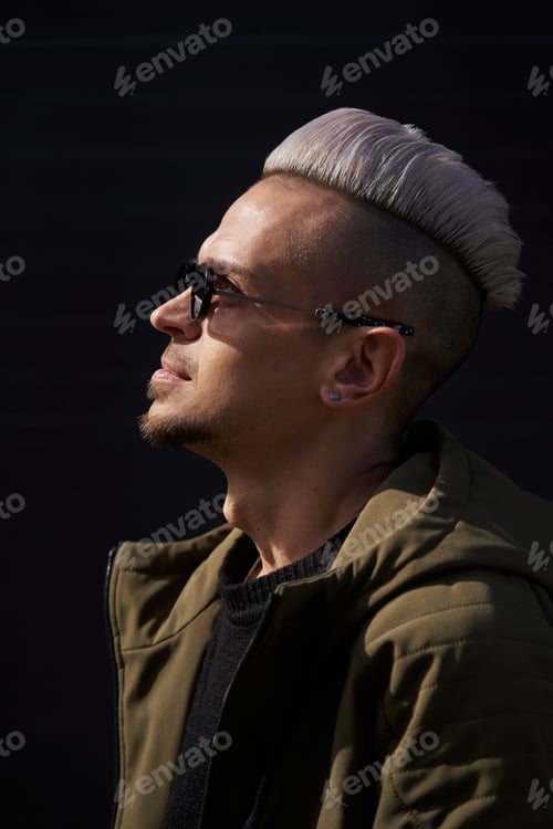 Preview: young caucasian man with white hair and sunglasses stands sideways in sunny color in a sweater and