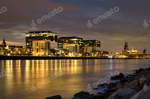 Preview: City buildings glowing near water at night in Cologne, Germany.