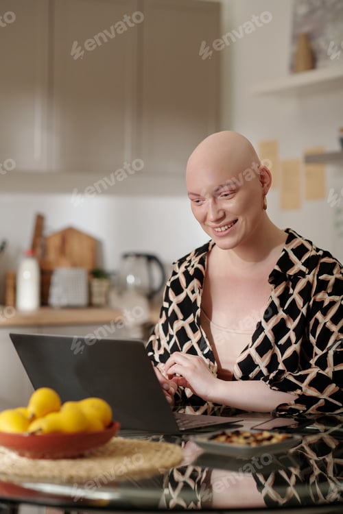 Preview: Happy young woman with bald head after chemotherapy using laptop