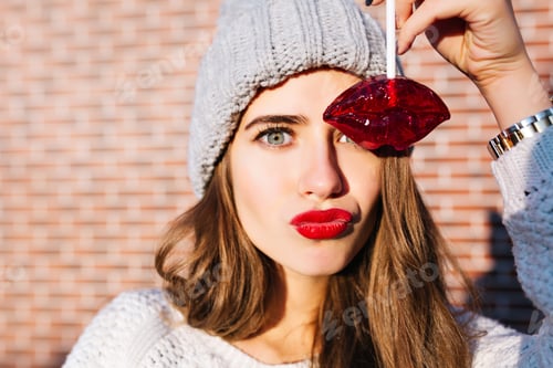 Preview: Closeup portrait pretty girl with long hair in knitted hat having fun with caramel red lips on wall