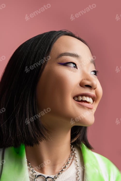 Preview: portrait of cheerful asian woman in metal necklaces and makeup looking away isolated on pink