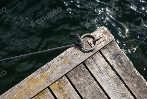 Preview: Old weathered wooden pier with a metal mooring ring and a stretched rope, against backdrop of sea