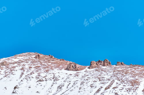 Preview: Rocks on top of mountain covered with snow over background of blue sky.