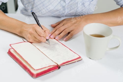Preview: The hand of a woman with a pen writes in her diary. Next on the table is a cup of tea