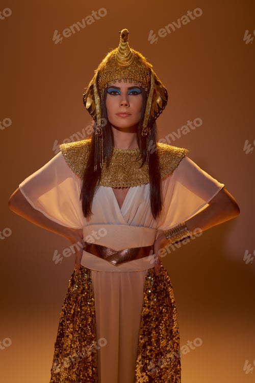 Preview: Stylish brunette woman in Egyptian headdress and look holding hands on hips on brown background