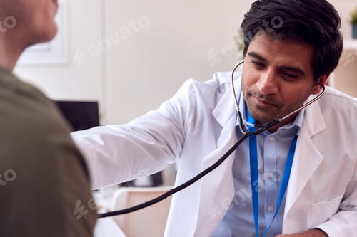 Preview: Male Doctor Or GP Wearing White Coat Examining Young Man Listening To Chest With Stethoscope