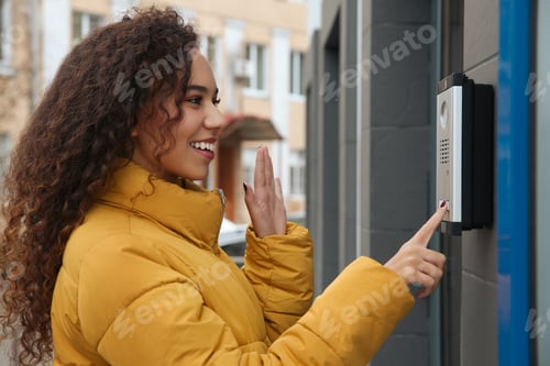 Preview: Young African-American woman ringing intercom while waving to camera near building entrance