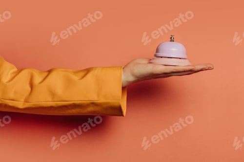 Preview: woman's hand in a yellow blazer holds a small bell against a pink background
