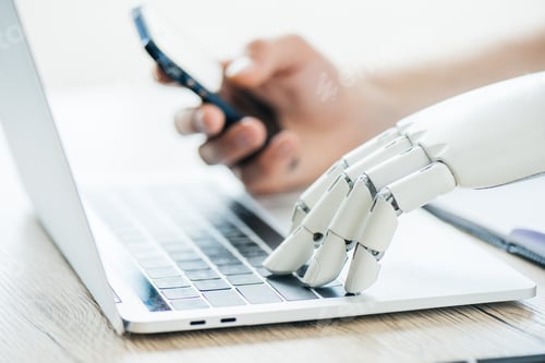 Preview: close-up view of human and robot hands using smartphone and laptop at wooden table