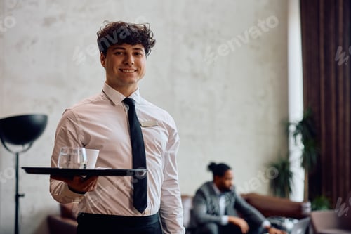 Preview: Happy waiter serving a guest in a hotel and looking at camera.