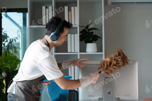 Preview: Man Cleaning Office Desk with Feather Duster and Headphones in Modern Home Office Environment