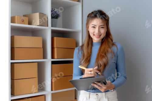Preview: Smiling young Asian business owner woman prepare parcel box and check online orders of product