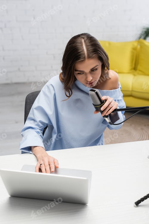 Preview: Brunette broadcaster in blue blouse talking at microphone and using blurred laptop while sitting