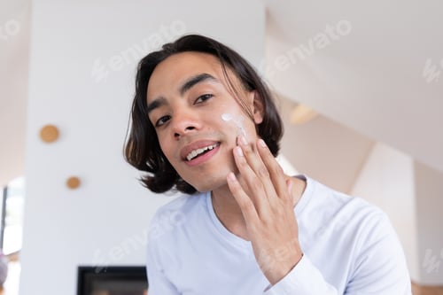 Preview: Happy biracial man looking in mirror, applying face cream in bathroom