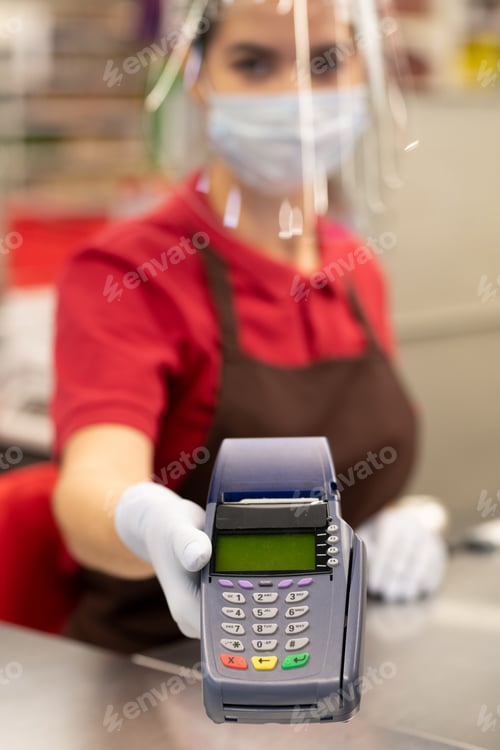 Preview: Gloved hand of cashier in protective screen and mask holding payment terminal