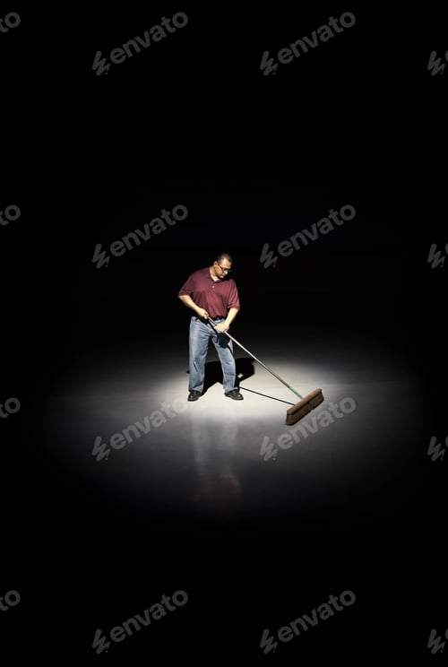 Preview: A man with a broom working in an area of a floor lit up by a spotlight.