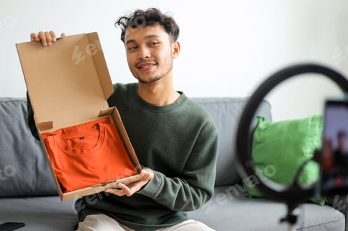 Preview: Young Asian Influencer Cheerfully Presenting Unboxing Orange T-Shirt To His Social Media Followers