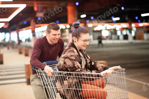 Preview: ✓ ↟ Cheerful happy young couple in casual clothes having fun and riding on a shopping cart