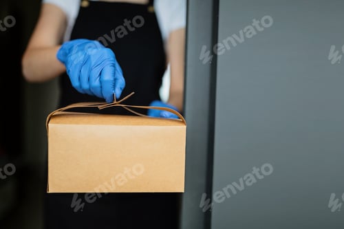 Preview: Cropped view of young waitress in latex gloves holding paper box package near window of cafe