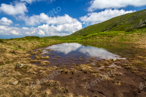 Preview: Blue sky with bright white clouds reflected in small lake between green hills on sunny day