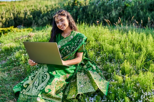 Preview: stylish and smiling young indian woman in sari using laptop while sitting on meadow in summer