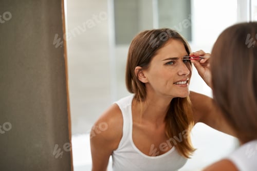 Preview: Shot of a young woman painfully tweezing her eyebrows in a bathroom mirror
