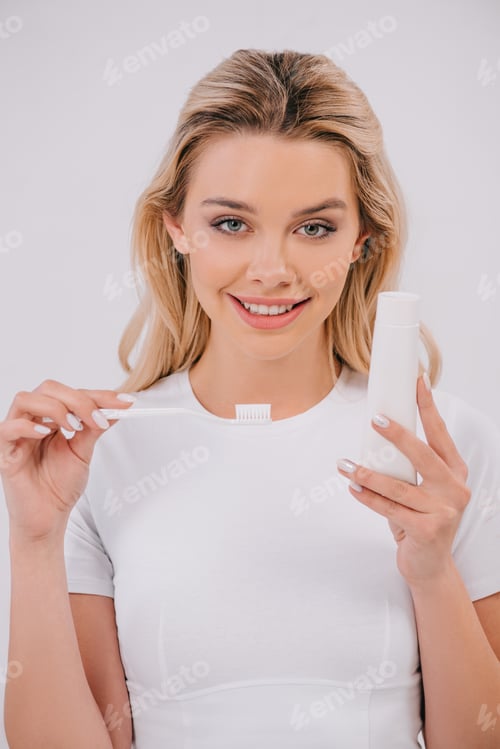 Preview: beautiful woman looking at camera while holding toothbrush and toothpaste with copy space isolated