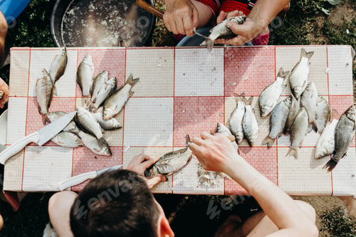 Preview: A young man cleans fish from scales while sitting at a table.
