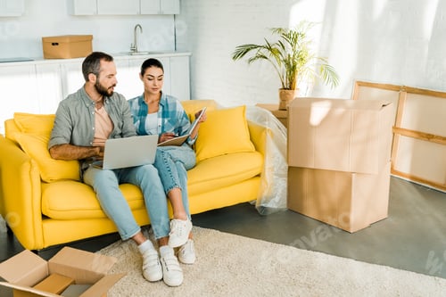 Preview: beautiful couple sitting on couch, using laptop and planning relocation to new house, moving concept
