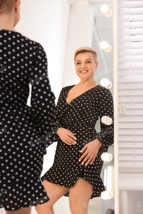 Preview: Beautiful mature woman looking at herself in large mirror with lights indoors