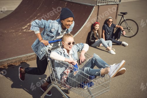 Preview: Boy carrying happy girl in shopping cart while friends sitting near ramp at skateboard park