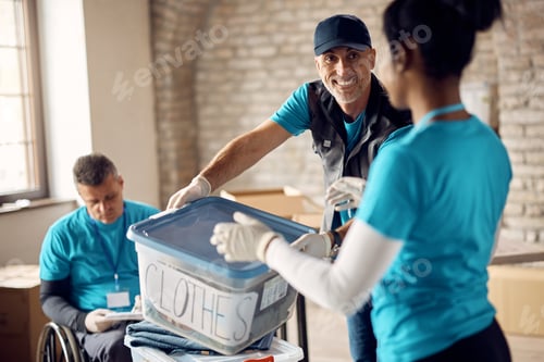 Preview: Happy mature volunteer passing box with packed clothes to his coworker at donation center.